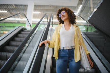 Kıvırcık saçlı, güneş gözlüklü, gülümseyen genç bir kadın. Süpermarkette yürüyen merdivende, yeniden kullanılabilir alışveriş çantasıyla.