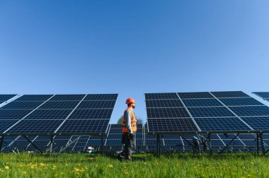 Güvenlik kaskı ve yelek giyen mühendis mavi gökyüzünün altındaki büyük bir güneş enerjisi santralinde yürüyor ve güneş panellerini inceliyor.