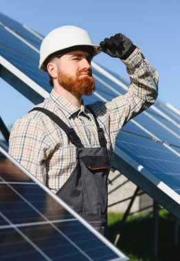 Sakallı mühendis, kaskını açık mavi gökyüzünün altında bir güneş enerjisi istasyonuna yerleştiriyor.