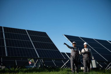İki işçi, bir Arap ve bir Avrupalı, açık mavi gökyüzü altında güneş panelleri kurmada işbirliği yaparak, takım çalışması ve yenilenebilir enerji çabaları sergiliyor