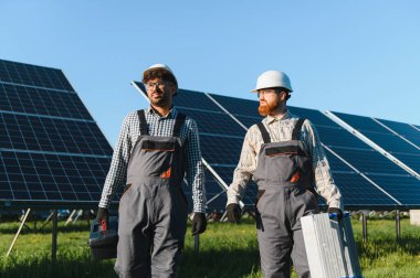 İki mühendis bir güneş enerjisi santralinde yürüyor, araçlar taşıyor ve sürdürülebilir enerji çözümlerine odaklanmış projelerde işbirliği yapıyorlar.