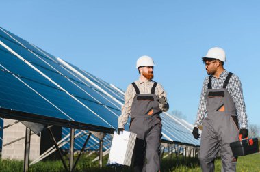 İki mühendis bir güneş enerjisi santralinde yürüyor ve tartışıyor, araçlar ve güneş panelleri taşıyor, yenilenebilir enerji ve takım çalışması sergiliyorlar.