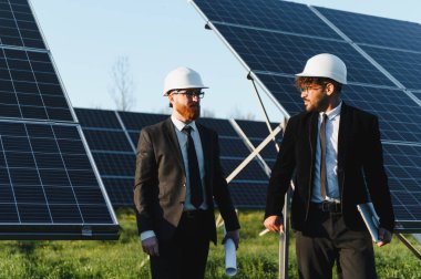 İki mühendis bir güneş enerjisi istasyonunda yürüyor ve tartışıyorlar, ellerinde planlar ve fotovoltaik panellerin performansını değerlendiriyorlar.