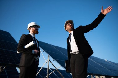 Güneş enerjisi santraline güneş panelleri kurulmasını tartışan iki mühendis, açık mavi gökyüzü altındaki gelecekteki gelişmelere işaret ediyorlar.