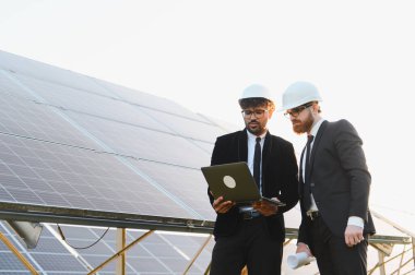 Kask ve giysili iki mühendis yenilenebilir enerji için güneş paneli çiftliğini analiz eden bir dizüstü bilgisayar kullanıyor.