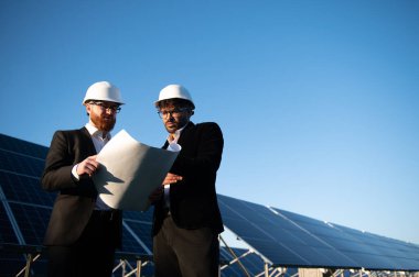 Şapkalı iki mühendis bir güneş enerjisi santralinin planlarını inceliyor, yenilenebilir enerji gelişimi ve işbirliği sergiliyorlar.