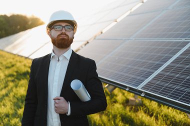 Yatırımcı sabit şapka ve gözlük takıyor, elinde bir plan tutuyor, sürdürülebilir enerji geliştirmeyi planlarken güneş paneli çiftliğini inceliyor.