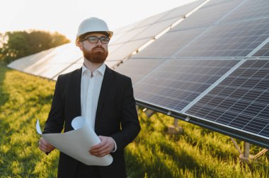 Yatırımcı sabit bir şapka takıyor ve elinde planlar tutuyor. Geniş bir güneş paneli çiftliğini inceliyor ve yenilenebilir enerji geliştirmeye odaklanıyor.