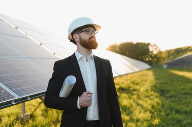 Güneş paneli çiftliğini gün batımında incelerken elinde bir planla, yenilenebilir enerji potansiyelini değerlendiren takım elbiseli bir yatırımcı.