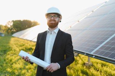 Yatırımcı sabit kasketli ve elinde bir planla güneş paneli çiftliğini incelerken gün batımında sürdürülebilir enerji çözümlerini değerlendiriyor.