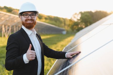 Bir güneş enerjisi istasyonundaki fotovoltaik panellere dokunurken, sürdürülebilir bir gelecek için yenilenebilir enerjiye yatırım yapan mühendis başparmaklarını gösteriyor