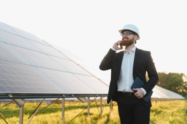 Güneş paneli çiftliğinde akıllı telefon kullanan tabletli ve beyaz şapkalı bir mühendis yenilenebilir enerjiyi tartışıyor.