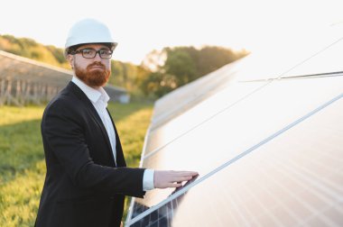Güneşli bir günde, büyük bir güneş çiftliğinde takım elbise ve şapka giyen yatırımcı güneş panelini teftiş ediyor.