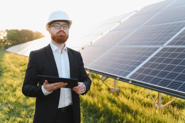 Güneş paneli çiftliğini gün batımında incelerken dijital tablet kullanan yatırımcı takım elbise giyiyor.