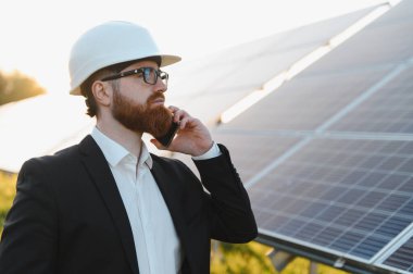 Fotovoltaik elektrik santralinde güneş panelleri yakınında akıllı telefon kullanan takım elbiseli bir yatırımcı.