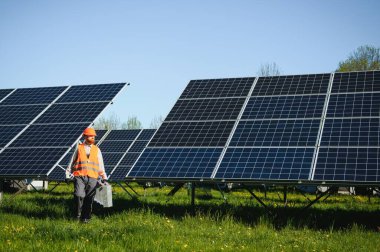 Güneşli bir günde güneş enerjisi istasyonunda çalışan mühendis fotovoltaik panellerin yanında yürüyen alet çantası taşıyor.