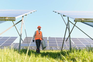Sürdürülebilir enerji tesisinde güneş paneli dizilerini denetleyen güvenlik ekipmanı bakım işçisi. Yeşil çimenler ve mavi gökyüzü endüstriyel güneş çiftliği çerçevesi