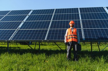 Güvenlik kaskı ve yeleği olan elektrik mühendisi fotovoltaik güneş panellerinin önünde yenilenebilir enerji ve sürdürülebilirliği temsil eden yeşil bir alanda duruyor.