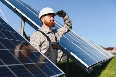 Beyaz kasklı ve tulumlu kızıl saçlı teknisyen, güneş enerjisi santralinde panellerin konumunu ayarlayan fotovoltaik paneller kuruyor.
