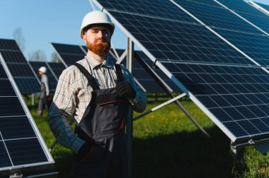 Güneşli bir günde güneş panelleri alanında poz veren tulum ve şapka takan sakallı mühendis.