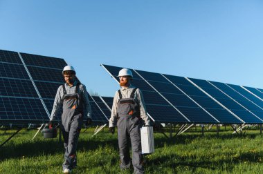 Güneş paneli çiftliğinde yürüyen iki mühendis, güneşli bir günde aletlerini taşıyor, bakım ve onarım yapıyorlar.