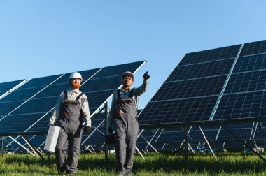 Güneş panellerinden oluşan geniş bir alanda yürüyen ve araçları taşıyan iki mühendis, bir fotovoltaik tesisi kuruyor ve koruyorlar.