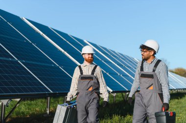 İki çoklu etnik mühendis güneş paneli çiftliğinde yürüyen ve aletleri taşıyan, tulumlu ve kasklı.