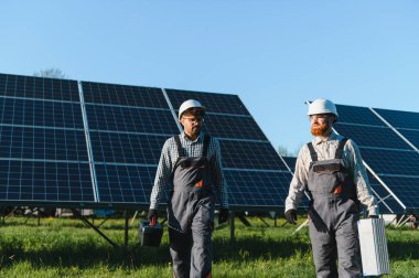 Güneş paneli çiftliğinde yürüyen iki mühendis, parlak bir güneşin altında araçları taşıyorlar, sürdürülebilir enerji çözümlerine odaklanıyorlar.