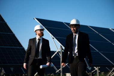 Güneş paneli çiftliğinde yürüyen iki mühendis proje detaylarını tartışıyor ve gelecek için sürdürülebilir enerji çözümleri üzerinde işbirliği yapıyorlar.