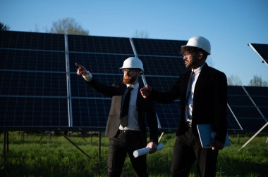Giysi ve başlık takan iki mühendis güneş paneli çiftliğindeki bir proje hakkında tartışıyorlar. Bir tanesi fotovoltaik modülleri işaret ediyor.