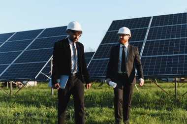 Güneş paneli çiftliğinde yürüyen ve tartışan takım elbiseli iki işadamı ellerinde dizüstü bilgisayar ve planlar tutuyorlar.