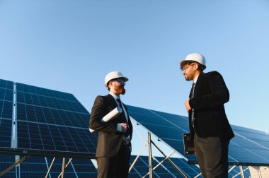 Güneş paneli çiftliğinde proje planlarını tartışan takım elbiseli iki mühendis yenilenebilir enerji ve sürdürülebilir kalkınma sergiliyorlar.