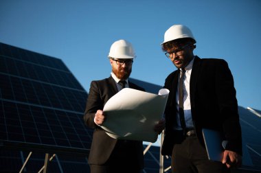 Kostümlü ve şapkalı iki mühendis, büyük bir güneş paneli kurulumunun önündeki planı inceliyor.