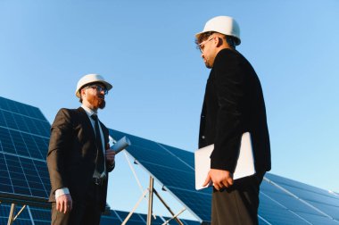 Takım elbiseli ve şapkalı iki işadamı, açık mavi bir gökyüzünün altında ellerinde planlar ve bir dizüstü bilgisayar olan bir güneş paneli kurulumunun yanında tartışıyorlar.