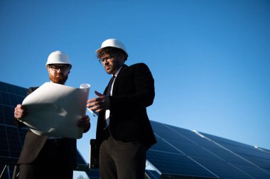 Kostüm ve şapka giyen iki mühendis, yenilenebilir enerji projesi üzerinde çalışan bir güneş paneli çiftliğinin planlarını tartışıyor.