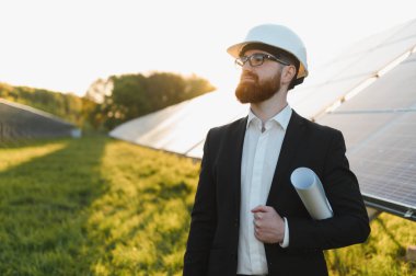 Mühendis bir şapka takıyor ve elinde bir plan tutuyor. Güneş paneli çiftliğini inceliyor. Gün batımının sıcacık parıltısıyla yıkanıyor.