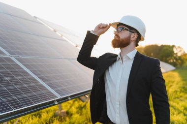 Sakallı iş adamı takım elbise ve koruyucu kask takıyor güneş enerjisi santralini gün batımında teftiş ediyor, kaskının güneşliğine dokunuyor.