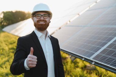 Yeşil enerji yatırımıyla tatmin olmuş fotovoltaik güneş panellerinin olduğu bir alanda başparmaklarını kaldıran bir mühendis.