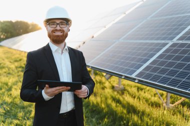 Yatırımcı başlık takıyor, elinde tablet tutarken gülümsüyor ve güneş paneli çiftliğinin gün batımının keyfini çıkarıyor.