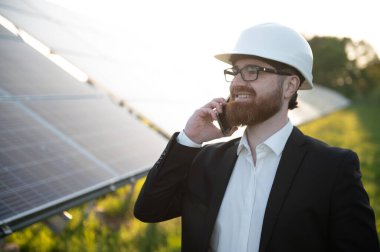 Güneş enerjisi istasyonundaki fotovoltaik panellerin yanında cep telefonu kullanarak takım elbise ve şapka takan gülümseyen yatırımcı