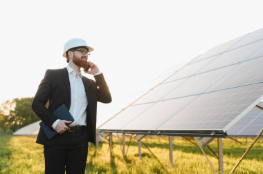 Yatırımcı telefonda konuşurken, elinde tablet tutarken büyük bir güneş paneli tesisini teftiş ediyor.