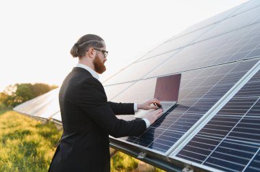 Bir elektrik santralindeki fotovoltaik güneş panellerini incelerken bir dizüstü bilgisayarda çalışan bir iş adamı, gün batımının güzelliğini yakalıyor.
