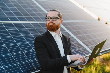 Dizüstü bilgisayar kullanan iş adamı, elektrik santralindeki fotovoltaik güneş panellerini denetliyor, sürdürülebilir kalkınma için yenilenebilir enerjiye yatırım yapıyor