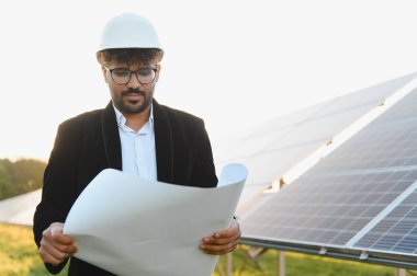 Arap mühendis başlık takıyor, güneş paneli çiftliğini inceliyor, elinde bir plan tutarken, açık havada yenilenebilir enerji geliştirmeye odaklanıyor.