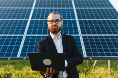 Kızıl saçlı işadamı gözlük takıyor, dizüstü bilgisayar tutuyor, güneş paneli çiftliğindeki yatırım olanaklarını ve büyüme potansiyelini analiz ediyor.