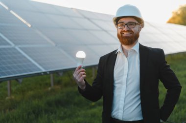 Sırıtkan işadamı, güneş panelleri önünde bir ampul tutarak, yenilenebilir enerji ve sürdürülebilir kalkınmayı destekliyor.