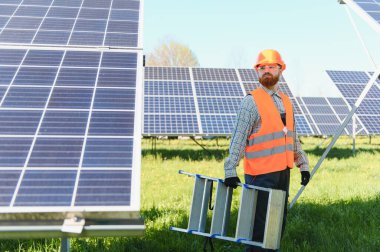 Güneş panellerini parlak, güneşli bir gökyüzünün altına yerleştirirken merdiven taşıyan erkek mühendis, yenilenebilir enerji çözümlerini teşvik ediyor.