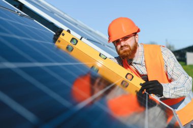 Güneş panellerini güneşli bir günde kurarken ruh seviyesini kullanan güvenlik yeleği ve kasklı erkek teknisyen yenilenebilir enerji ve sürdürülebilir uygulamaları teşvik ediyor.