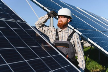 Erkek mühendis, güneş enerjisi santralinde çalışırken kaskını ayarlıyor ve yenilenebilir enerji üretiminin güvenliğini ve verimliliğini sağlıyor.