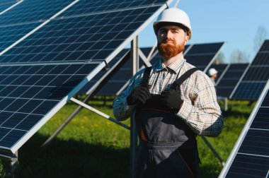 Kızıl kafalı mühendis, büyük bir güneş enerjisi santraline fotovoltaik paneller yerleştirirken iş kıyafetlerini ayarlıyor.